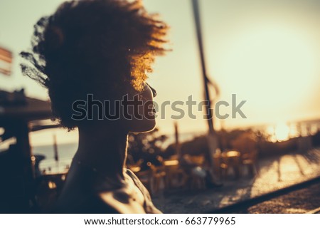 Silhouette of young Brazilian girl with curly afro hair illuminated by orange light looking on coastline while standing on embankment on windy summer evening, with beach cafe in defocused background