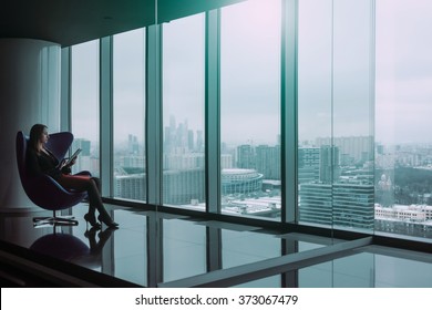 Silhouette of young business woman with tablet seated on a chair, winter city landscape outside the window on the background