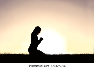 A Silhouette Of A Young Christian Woman Kneeling In Prayer On A Summer Day.