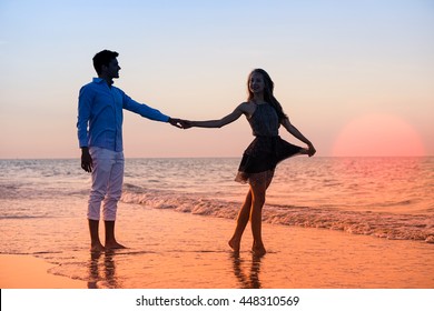 Silhouette Of Young Couple Walking At Sunset On The Beach - Happy Lovers Holding Hands At Night By The Ocean With Big Red Sun Background - Iconic Concept Of Love And Tropical Holidays With Warm Colors