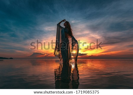silhouette of young fashionable woman standing in water on the beach at sunset
