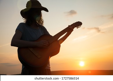 silhouette of young free woman in straw hat playing country music on a guitar at sunset, copy space