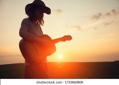 Silhouette Of Young Free Woman In Straw Hat Playing Country Music On A Guitar At Sunset, Copy Space