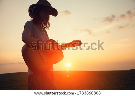 silhouette of young free woman in straw hat playing country music on a guitar at sunset, copy space