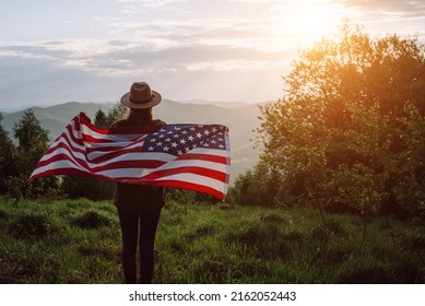 Silhouette of young girl in hat holding American Flag looking out at landscape. Patriotic american woman 20s old years enjoying nature during summer weekend. July 4th forth independence day concept