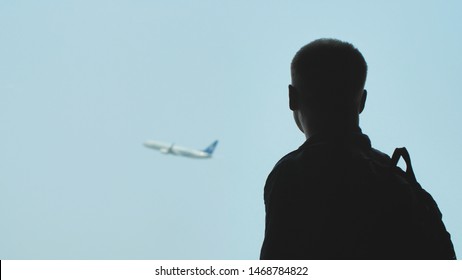 Silhouette Of A Young Man With A Backpack On His Back Watching The Airplane Take Off Against The Blue Sky.