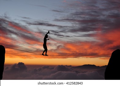 Silhouette of young man balancing on slackline high above clouds and mountains during sunset. Slackliner balancing on tightrope between two rocks sun and clouds behind, highline silhouette. - Powered by Shutterstock