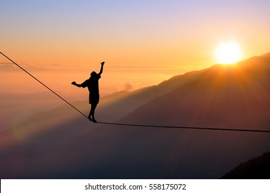 Silhouette of young man balancing on slackline high above clouds and mountains, sun, beautiful colorful sky and clouds behind. Slackliner balancing on tightrope between two rocks, highline silhouette. - Powered by Shutterstock