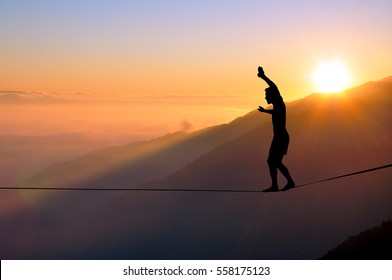 Silhouette of young man balancing on slackline high above clouds and mountains, sun, beautiful colorful sky and clouds behind. Slackliner balancing on tightrope between two rocks, highline silhouette. - Shutterstock ID 558175123