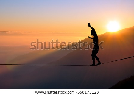 Silhouette of young man balancing on slackline high above clouds and mountains, sun, beautiful colorful sky and clouds behind. Slackliner balancing on tightrope between two rocks, highline silhouette.