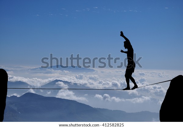 Silhouette of young man balancing on\
slackline high above clouds and mountains. Slackliner balancing on\
tightrope between two rocks, highline\
silhouette.