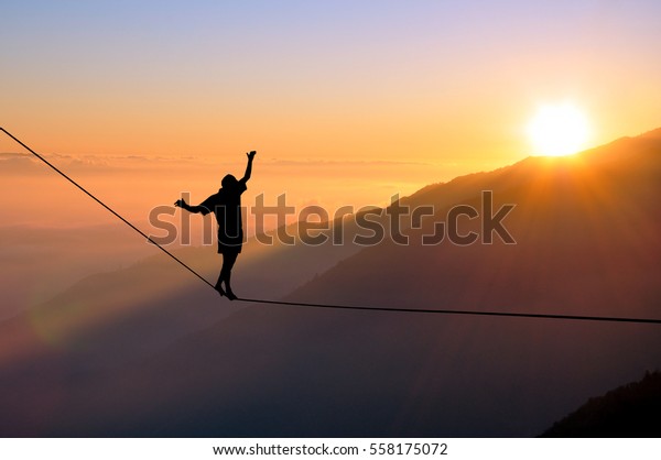 Silhouette of young man balancing on\
slackline high above clouds and mountains, sun, beautiful colorful\
sky and clouds behind. Slackliner balancing on tightrope between\
two rocks, highline\
silhouette.