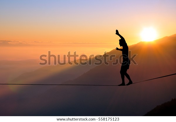 Silhouette of young man balancing on\
slackline high above clouds and mountains, sun, beautiful colorful\
sky and clouds behind. Slackliner balancing on tightrope between\
two rocks, highline\
silhouette.