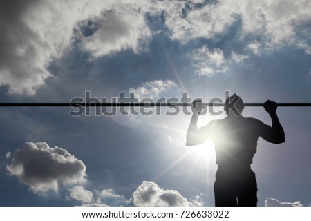 Silhouette of a young man doing pull-up exercise on a horizontal bar against a blue cloudy sky with sun flare.
