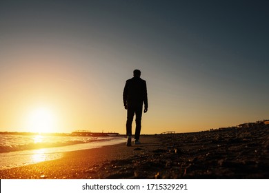 Silhouette of young man on the beach in Italy. Back view silhouette of a walking man running along on the beach at sunset with sun in the background. Man walking on the beach looking on sea at sunset.