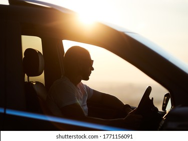 Silhouette Of A Young Man Sitting In His Car, Wearing Sunglasses And Looking At The Sunset, Hand On The Helm, Bright Sun On The Car Roof, Close Up, Side View