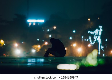 Silhouette Of A Young Man Sitting Outdoor On The Ground Alone At Night, Looking Sad At Lam Vien Square Park. Blurred Neon Lights From Background Creating Beautiful Bokeh And The Surreal Aesthetic