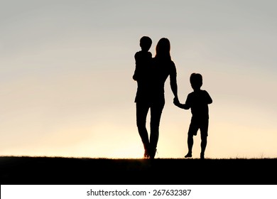 Silhouette Of A Young Mother Walking And Lovingly Holding Hands With Her Happy Little Child, While Holding His Baby Brother, Outside In Front Of A Sunset In The Sky.