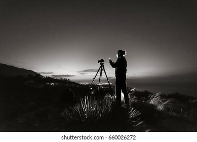 Silhouette of young photographer looking into his cell phone and making panoramic photo of a night city with camera on tripod. Black and white photography.