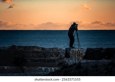 Silhouette of young photographer in twilight capturing sunset at dusk with orange cloudy sky by the ocean. professional landscape photography with tripod and camera. - Powered by Shutterstock - Get 15% off with code: PIKWIZARD15