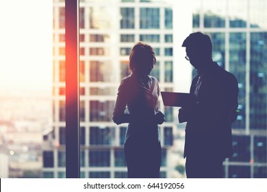 Silhouette of young purposeful two business partners working on digital tablet while standing near skyscraper office window,man and woman professional bankers using touch pad for teamwork during break - Powered by Shutterstock