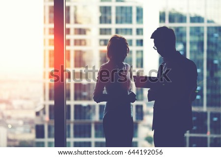 Silhouette of young purposeful two business partners working on digital tablet while standing near skyscraper office window,man and woman professional bankers using touch pad for teamwork during break