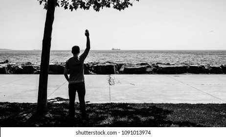 Silhouette Of Young Sad Man Or Boy Standing Alone At Seaside, Waving His Hand To Distant Ship. Male Feeling Lonely / Sorry. Time To Go. Missing Someone. Desire To Go Far Away. Time To Say Goodbye.