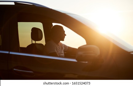 Silhouette Of A Young Serious Man Sitting In His Car, Wearing Sunglasses And Looking At The Sunset, Bright Sun On The Car, Close Up, Side View