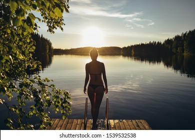 Silhouette of young shapely woman in bikini stand at pier after sauna. Summer evening sunset on the lake in Finland. Calm finnish nature. - Powered by Shutterstock