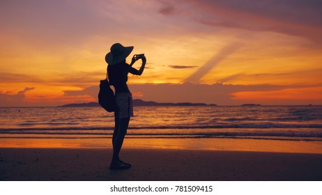 Silhouette of young tourist woman in hat taking photo with cellphone during sunset in ocean beach - Powered by Shutterstock