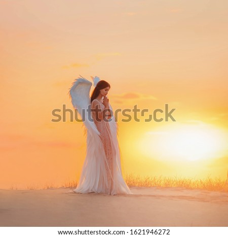 Silhouette young woman angel stands in desert praying. Creative glamour design costume clothes with bird wings feathers. Bright yellow color sunset dramatic heaven. Photo Shoot Divine Fairy Spirit