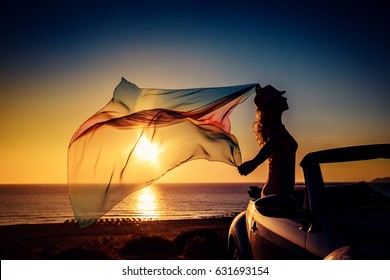 Silhouette Of Young Woman At The Beach. Girl With Flying Fabric Against Sunset Sky And Sea Background. Female Traveling By Car. Summer Vacation And Travel Concept