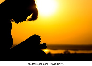 silhouette of young woman bowed her head and praying to God in the nature, the girl holds her hands on the Bible near the tree at sunset, the concept of religion and spirituality