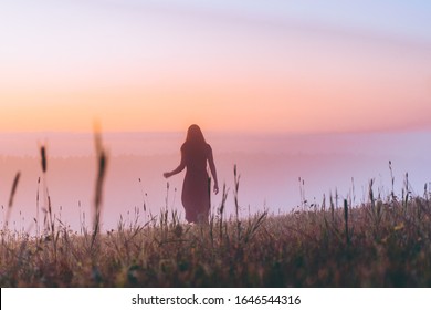 silhouette of young woman in a dress walking on a hill on sunrise. female running on a foggy field and enjoying forest landscape. adventure, freedom, morning, nature concept. dew on grass.