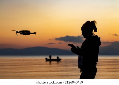 silhouette of young woman flying drone at sunset. Sea and fishing boat in the background. drone and woman taken in reverse light.