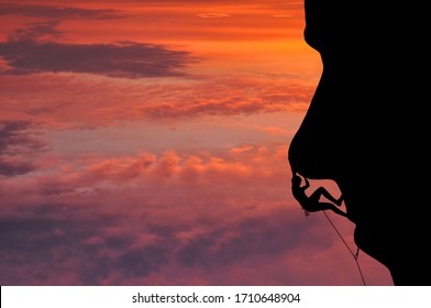 Silhouette Of Young Woman Lead Climbing The Nose On Cliff In The Shape Of Human Face. Abstract View Of Overhanging Cliff, Beautiful Colorful Sky And Clouds. Climber On Top Rope, Hanging On Rock.