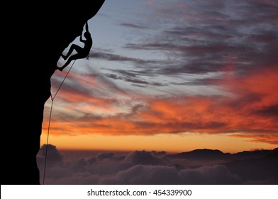 Silhouette of young woman lead climbing on overhanging cliff high above clouds and mountains, sun, beautiful colorful sky and clouds behind. Climber on top rope, hanging on rock during sunset.