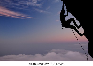 Silhouette Of Young Woman Lead Climbing On Overhanging Cliff High Above Clouds And Mountains, Sun, Beautiful Colorful Sky And Clouds Behind. Climber On Top Rope, Hanging On Rock.