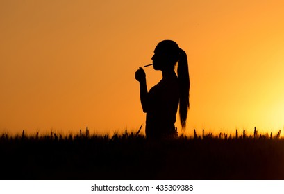 Silhouette of young woman lighting cigarette at sunset in wheat field - Powered by Shutterstock