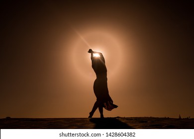 silhouette of young woman with long skirt dancing in evocative and magic way on top of desert dune at sunset with sun high in the sky
