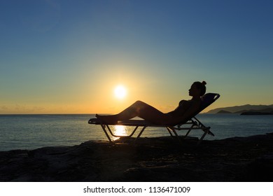 Silhouette of the young woman is lying on the deckchair at sunset.