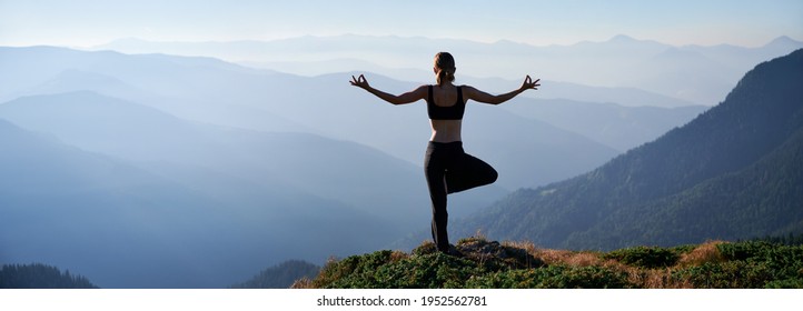 Silhouette of young woman performing yoga pose on grassy hill and looking at beautiful mountains. Sporty woman standing on one leg while practicing yoga outdoors in the morning.