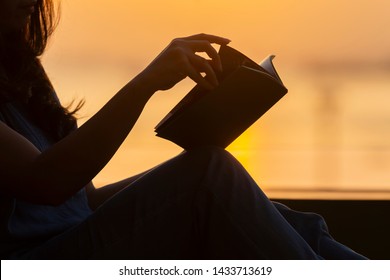 Silhouette Of A Young Woman Reading Book At Sunset. Calm And Warm Tone 