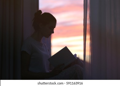 Silhouette Of Young Woman Reading Book On Windowsill At Home