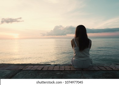 Silhouette of young woman sitting alone on back side outdoor at tropical island beach missing boyfriend and family in summer sunset. Sad and lonely concept in dark and vintage tone.