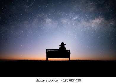 Silhouette Of Young Woman Sitting On Old Wooden Chair Looking At The Night Sky And Looking At The Beauty Of The Stars And The Milky Way Alone On Top Of The Mountain.