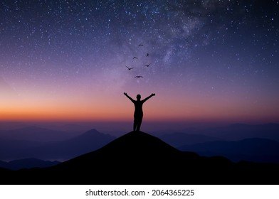 Silhouette of young woman standing alone on top of mountain and raise both arms praying and free bird enjoying nature on beautiful night sky, star, milky way background. Demonstrates hope and freedom.