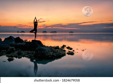 Silhouette of a young woman standing on the rocks by the sea in a yoga pose against the backdrop of a spectacular sunset and a huge rising full moon on a summer evening - Powered by Shutterstock