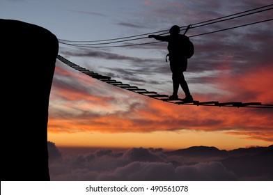 Silhouette of young woman walking to the cliff on the via ferrata bridge high above clouds and mountains, sun, beautiful colorful sky and clouds behind. Climber on via ferrata bridge during sunset.