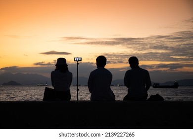 Silhouette Of Youngsters Students Boy And Girl Chat And Have Fun And Take Selfie Of Mobile Phone Camera During Sunset In West Kowloon Waterfront Promenade, Hong Kong In Evening. Back Light Shot